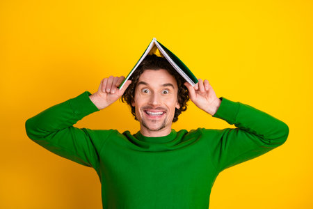 Cheerful young man posing playfully with a book in a green sweater on a vibrant yellow background