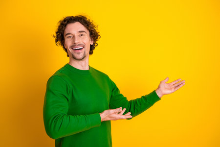 Young man with curly hair in a green sweater gesturing confidently against a yellow background, cheerful expressionの写真素材