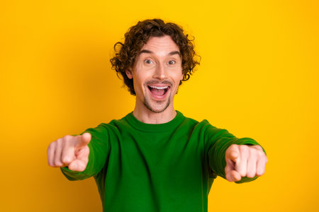 Cheerful young man in green sweater gesturing towards camera against yellow backgroundの写真素材