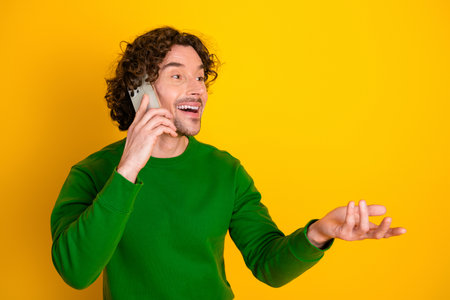 Happy young man talking on a smartphone against a vibrant yellow background while wearing a green sweaterの写真素材