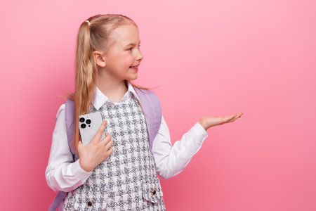 Smiling schoolgirl holding smartphone and gesturing while standing against pink background in school...
