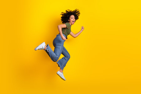 Happy young woman in casual wear jumping against yellow background, expressing joy, energy, and a...