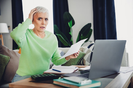 Mature woman in bright sweater using laptop and reading documents while sitting on sofa in modern...