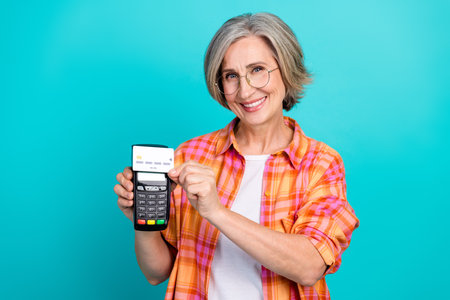 Elderly Woman Smiling and Demonstrating a Payment Terminal with a Credit Card on a Vibrant Turquoise Backgroundの写真素材