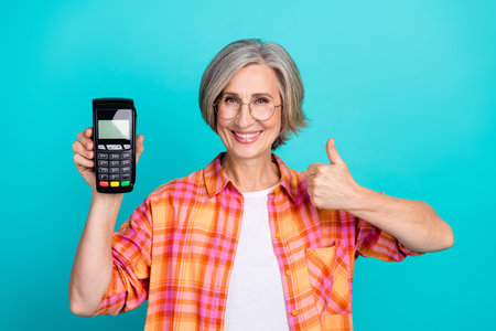 Happy woman in casual checkered shirt holding payment terminal against teal background with a thumbs-up gestureの写真素材