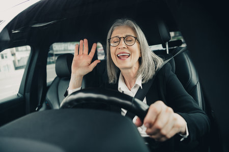 Mature professional woman driving a car while smiling and waving in a modern urban settingの写真素材