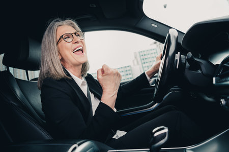 Mature senior businesswoman driving a car celebrating success wearing stylish formalwear in an urban daytime environmentの写真素材