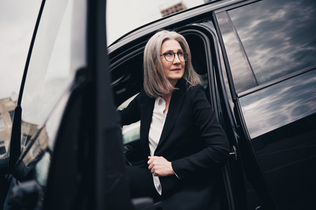 Confident mature businesswoman stepping out of a car in an urban setting, dressed in formal wear, exuding professionalism and eleganceの写真素材