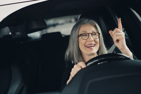 Confident mature woman with gray hair driving a car in the city on a bright day, smiling and...