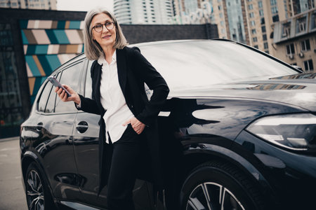 Successful mature businesswoman with grey hair and elegant outfit standing next to a car in an urban street settingの写真素材