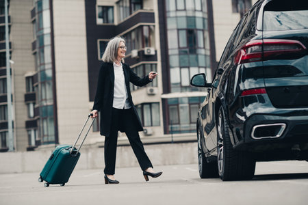 Confident businesswoman with grey hair walking outdoors with luggage near a stylish car in an urban city environmentの写真素材