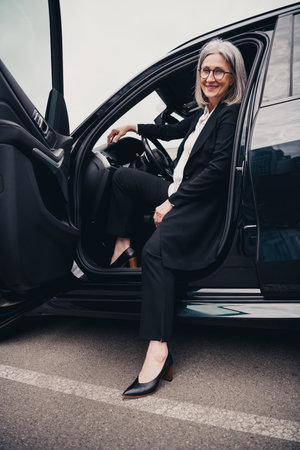 Elegant mature businesswoman sitting in car doorway outdoors, dressed in elegant formalwear, enjoying a productive day.の写真素材