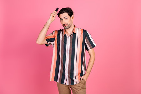 Confident young man combing his hair while standing against a pink background embracing a playful and stylish vibeの写真素材