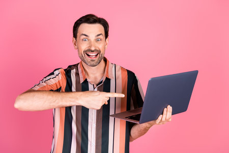 Excited man in striped shirt using a laptop against a pink background, gesturing happily and showcasing an energetic demeanorの写真素材