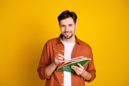Young man with beard holding notebooks and pencil standing against a bright yellow backgroundの写真素材