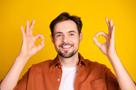 Smiling young man in casual orange shirt making an OK gesture against vibrant yellow background, symbolizing positivityの写真素材