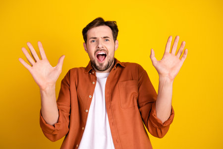 Excited young man in an orange shirt posing with raised hands against a vibrant yellow background, expressing excitementの写真素材