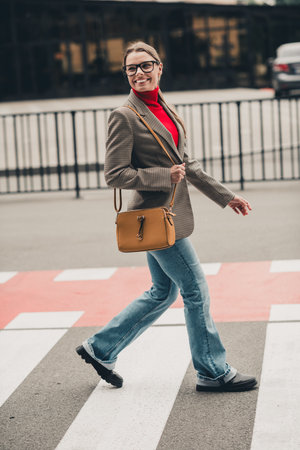 Young professional businesswoman crossing urban street wearing formal outfit including blazer and jeans smiling confidentlyの写真素材
