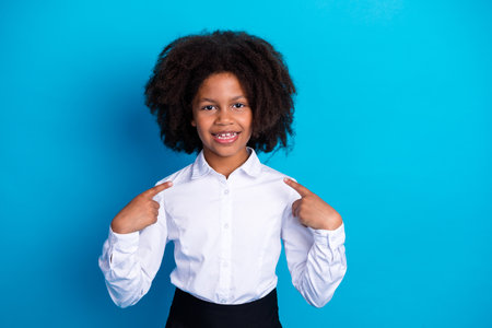 Cheerful young girl pointing at herself against blue background wearing white shirt emphasizing self-confidence and joyの写真素材