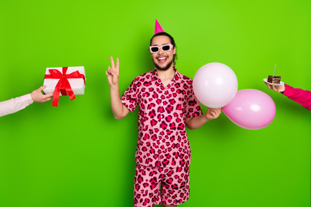 Cheerful young man in vibrant outfit celebrating with balloons and a gift against green festive...