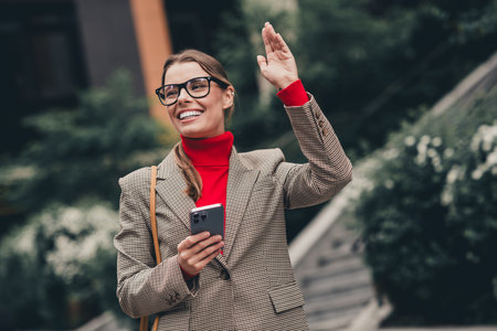 Confident businesswoman waving while holding phone outdoors in urban setting, wearing formal attire and red sweaterの写真素材