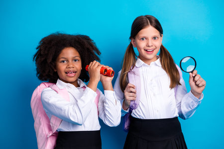 Two cheerful schoolgirls holding a magnifying glass and binoculars against a solid blue background, promoting curiosity and educationの写真素材
