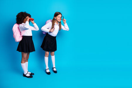 Two schoolgirls happily exploring with binoculars and magnifying glass against a bright blue backgroundの写真素材