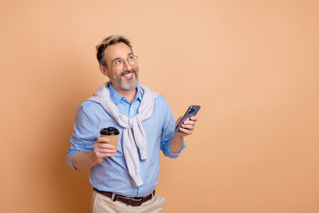 Elderly stylish man holding coffee and mobile phone with beige background representing modern...