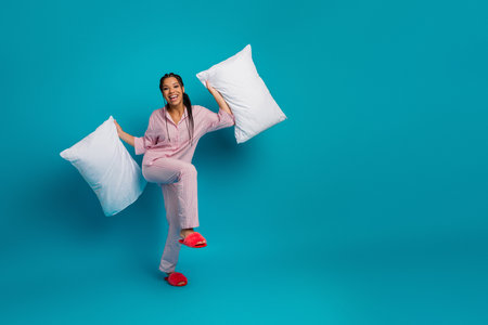 Cheerful young woman in pink pajama holding white pillows on a blue background representing fun and relaxationの写真素材
