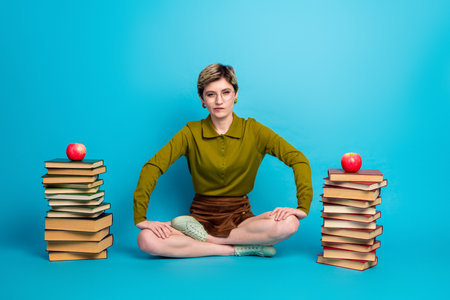 Young trendy woman seated on floor with two stacks of book against a vibrant blue background, promoting education and styleの写真素材