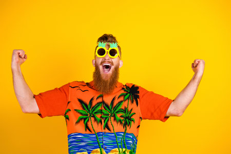 Cheerful young man in tropical print t-shirt celebrating against vibrant yellow background wearing pineapple sunglassの写真素材