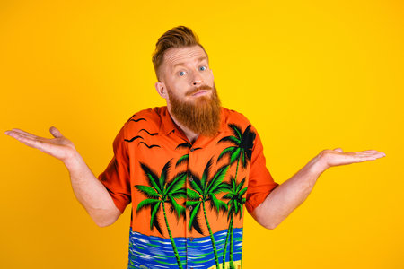 Young bearded man in tropical printed shirt gesturing casually against vibrant yellow background,...