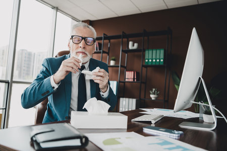 Senior businessman in office setting holding tissues while working at desk with computer emphasizing workspace environmentの写真素材