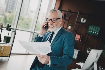 Mature businessman in modern office holding documents while talking on phone, dressed in formalwear, professional workの写真素材