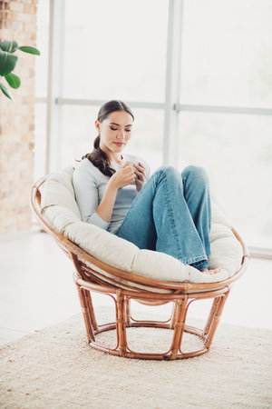 Young woman relaxing on a cozy rattan chair in a modern bright living room with natural light, enjoying a cup of coffeeの写真素材