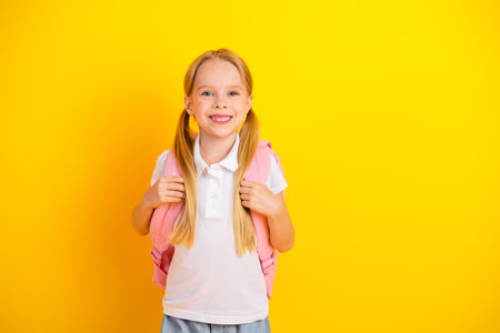 Young schoolgirl with backpack smiling on yellow background showcasing happiness, youth, primary...