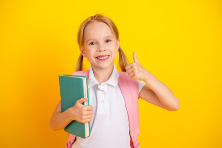 Happy schoolgirl with backpack holding a book and showing a thumbs up gesture with a bright yellow backgroundの写真素材