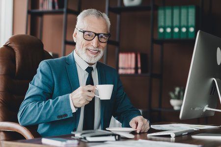 Confident Mature Businessman at Office Workspace Holding Coffee and Working on Computer in Professional Atmosphereの写真素材