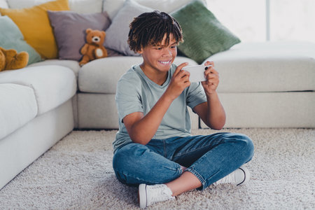 Young boy happily playing on his smartphone while sitting on a cozy floor in a modern and comfortable living roomの写真素材