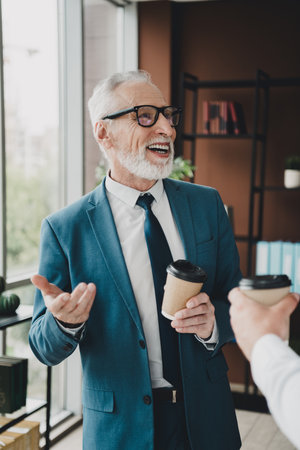 Confident senior businessman in formal suit enjoys coffee in modern office environment, showcasing leadership and positivityの写真素材