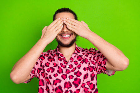 Young man in a vibrant pink leopard print shirt smiling and covering his eyes playfully against a vivid green background.の写真素材