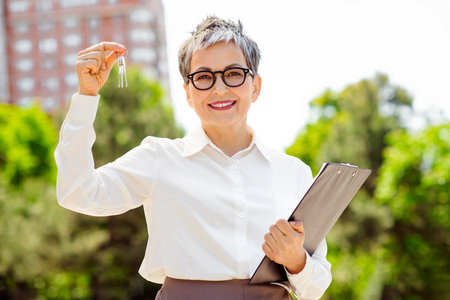 Mature professional businesswoman displaying keys outdoors near urban buildings in a formal white shirt holding a clipboard