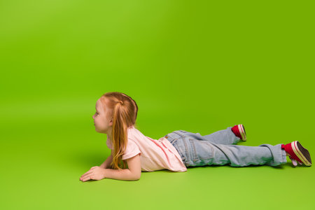 Charming little schoolgirl lying on the ground against a vibrant green background, expressing joy, innocence, and positive energyの写真素材