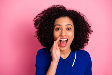 Excited young woman with curly hair smiling and shouting with hand to mouth against pink background