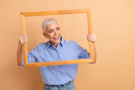 Cheerful Mature Woman Holding a Frame, Expressive and Stylish Portrait on a Neutral Beige Backgroundの写真素材