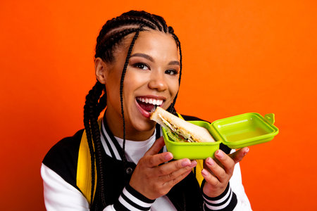 Smiling student enjoys lunch while holding a sandwich in a container against vibrant orange backgroundの写真素材