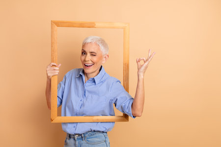 Confident mature woman with short white hair posing with a wooden frame on a beige background,...