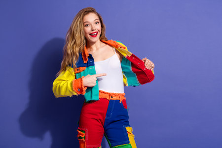 Young cheerful woman in vibrant patchwork outfit confidently posing against a purple backgroundの写真素材