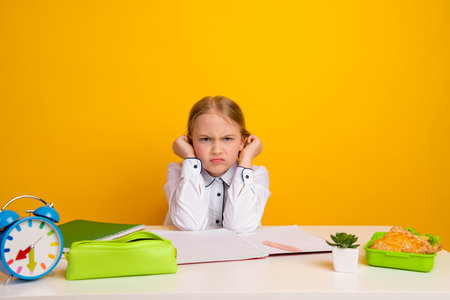 Upset schoolgirl in a white uniform sitting at desk with study items and food against vibrant yellow...