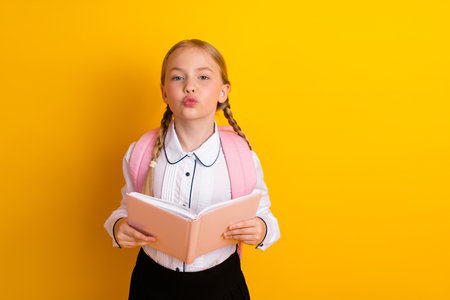 Enthusiastic schoolgirl holding a book and wearing a backpack against a vibrant yellow background,...